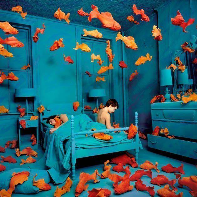 a blue hued bedroom with a child on the bed filled with orange goldfish in the air and on the floor