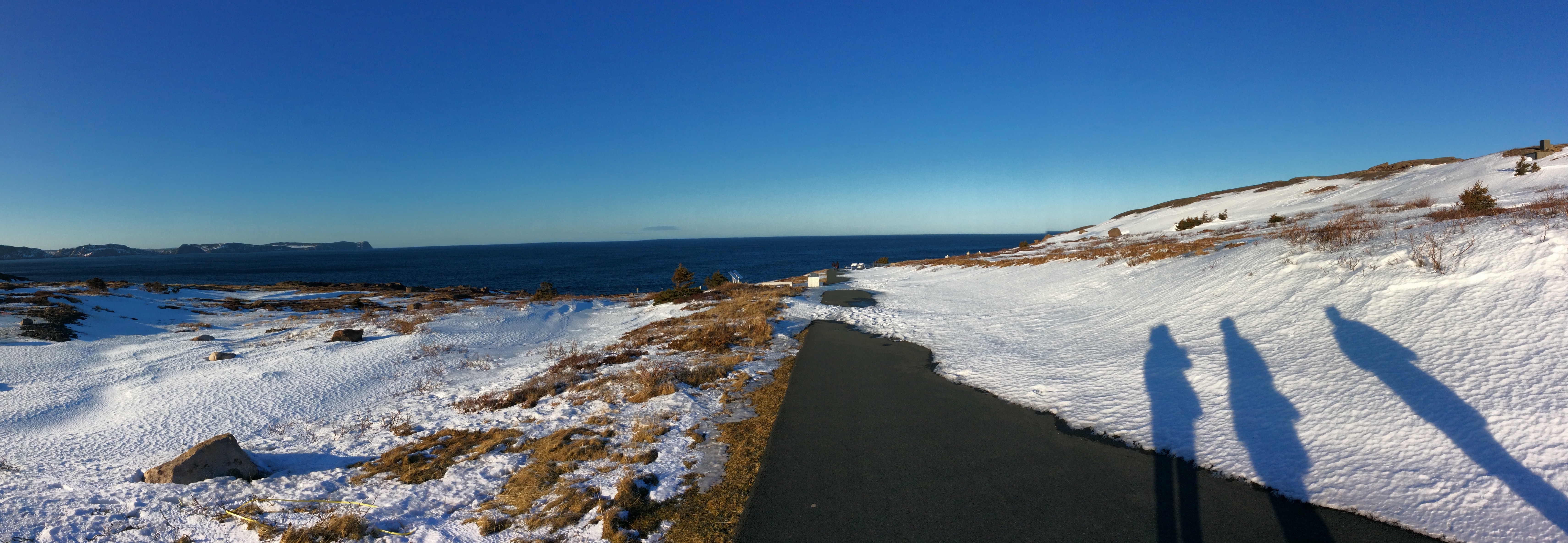 shadows of three people on a batch of snow with a dark path ahead of them, sea in the background and a blue sky above. 