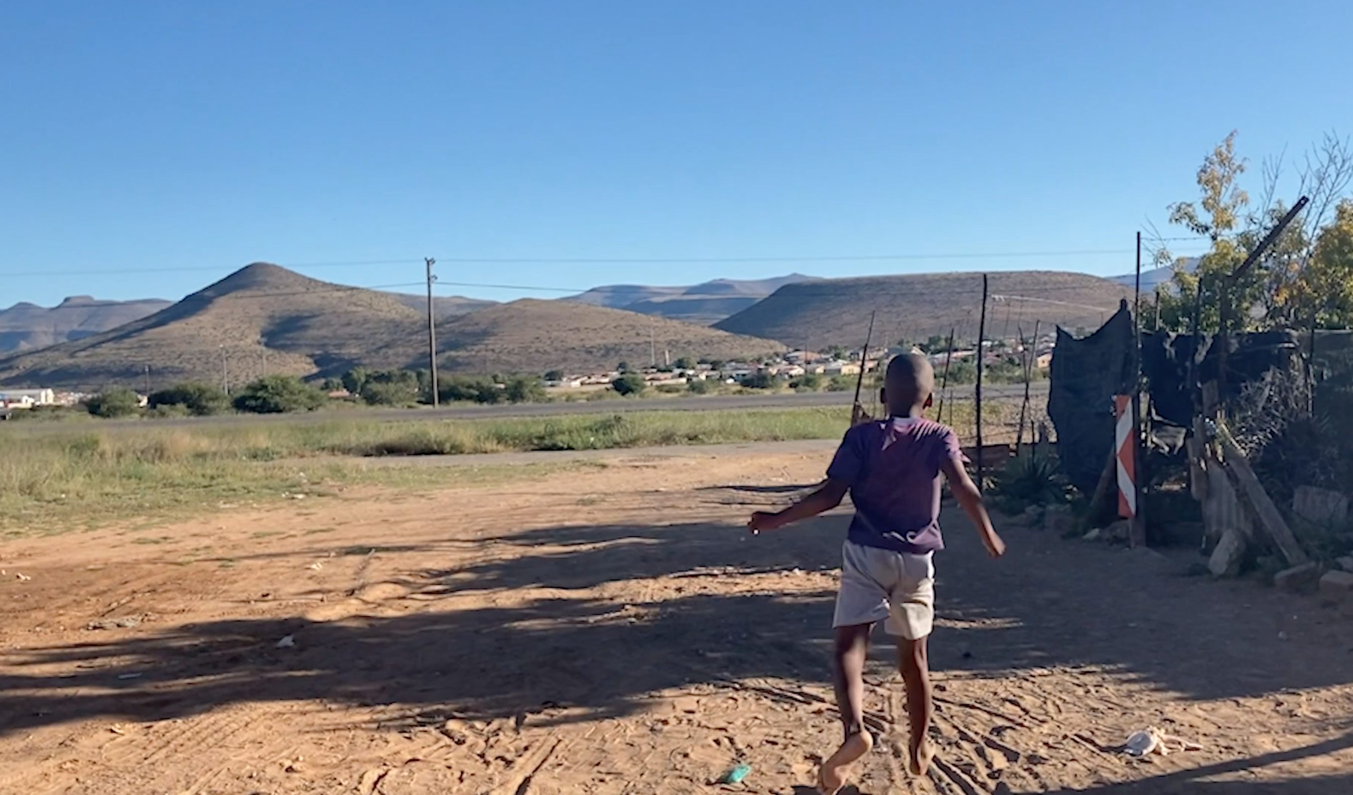 A barefoot boy runs on a sandy road. There are hills in the background.