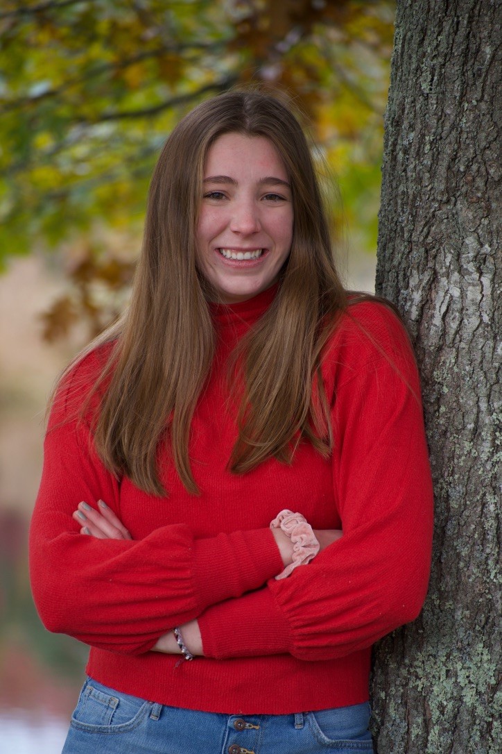 Faye Parker is facing the camera wearing a red sweater, arms crossed, leaning against a tree.