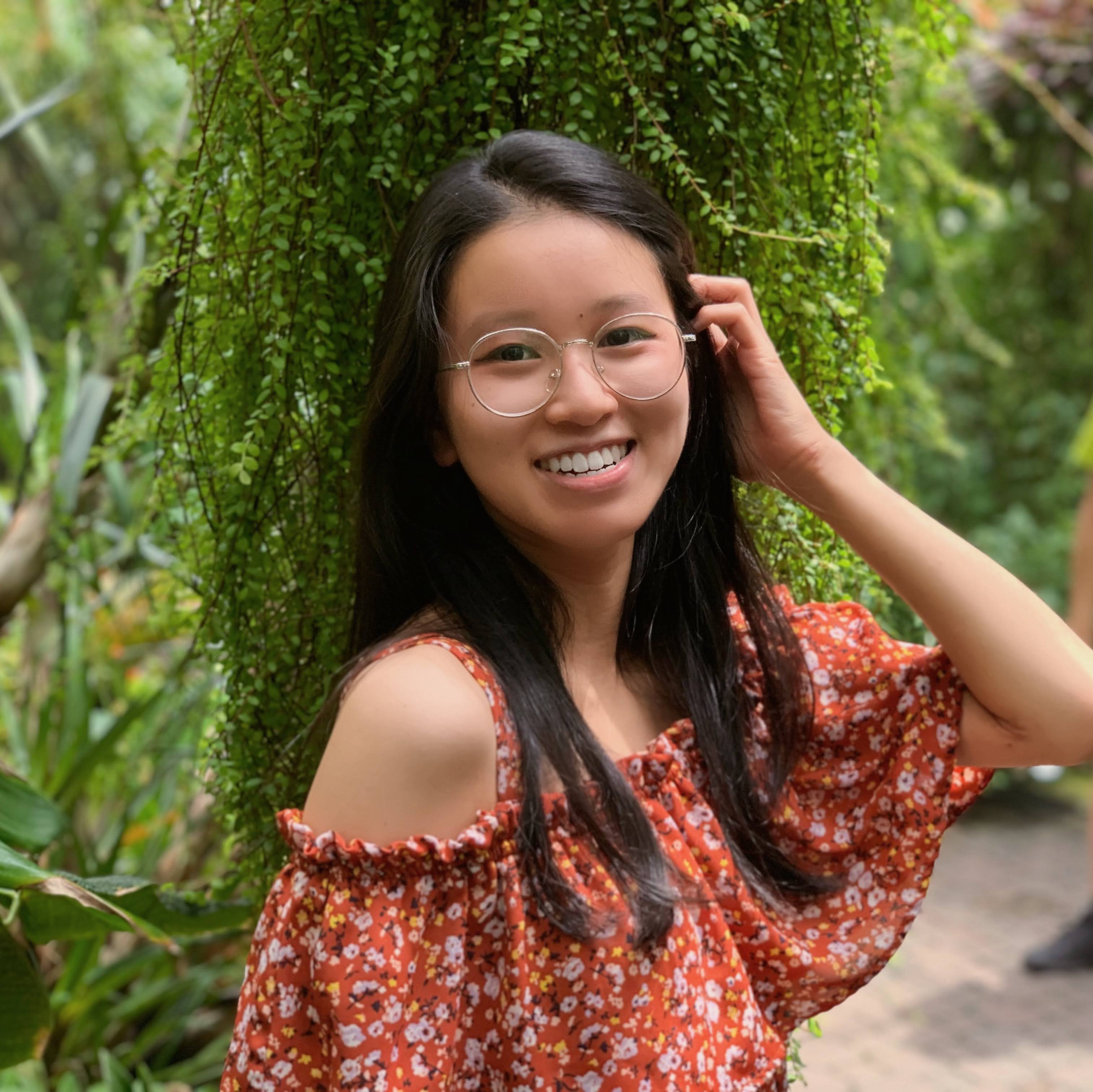 Mary Tuyetnhi Tran in an orange patterned shirt turns to face the camera, with their hand tucking hair behind their ear. They are in front of lush green foliage. 