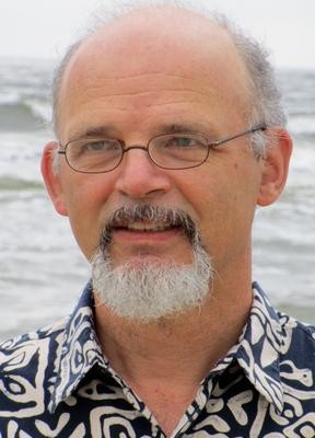 Portrait of Simon Richter. Simon has short gray hair, facial hair, and wire rimmed glasses. They are standing in front of a body of water. 