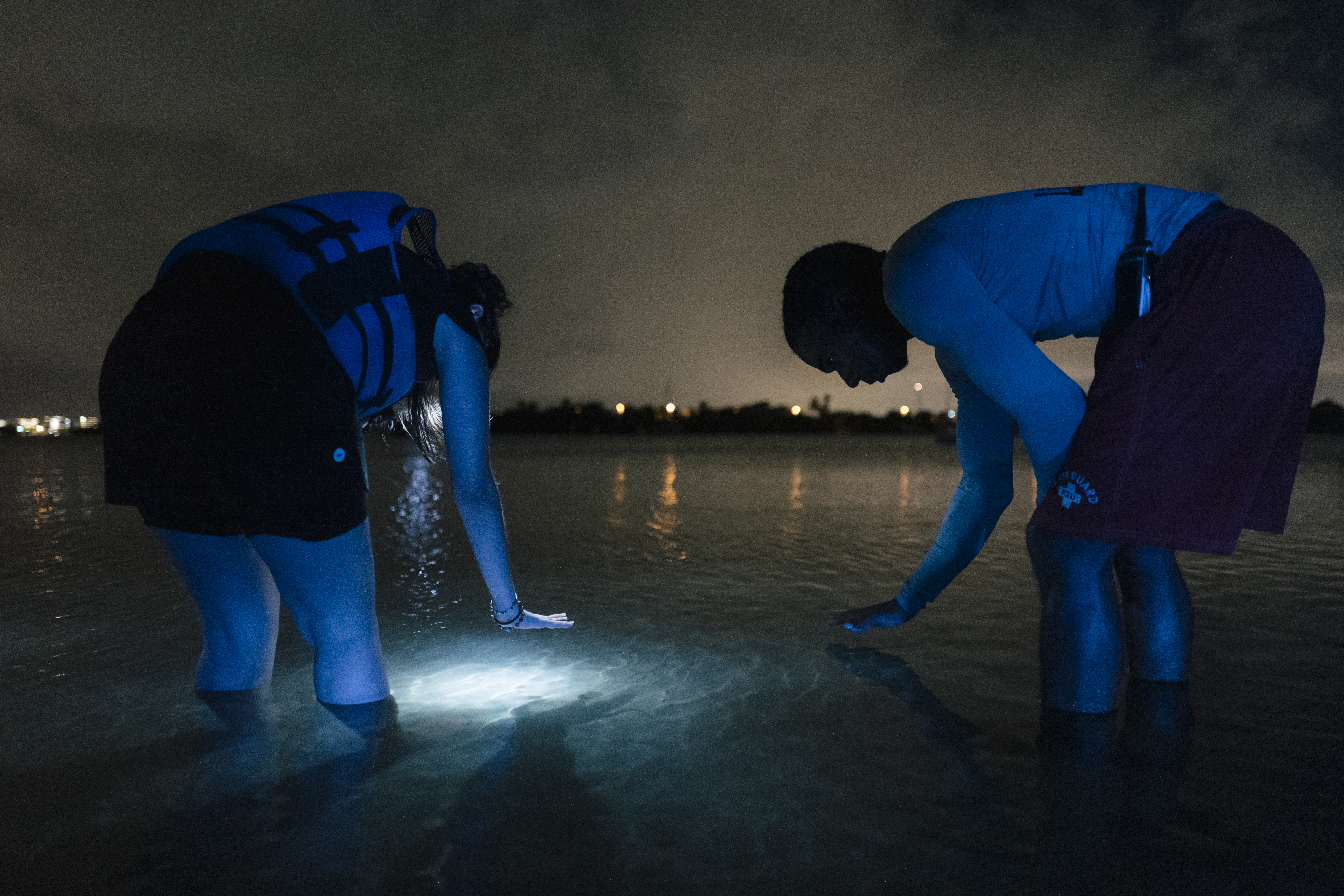 two people bent close to the water with flashlights in the dark peer into the river