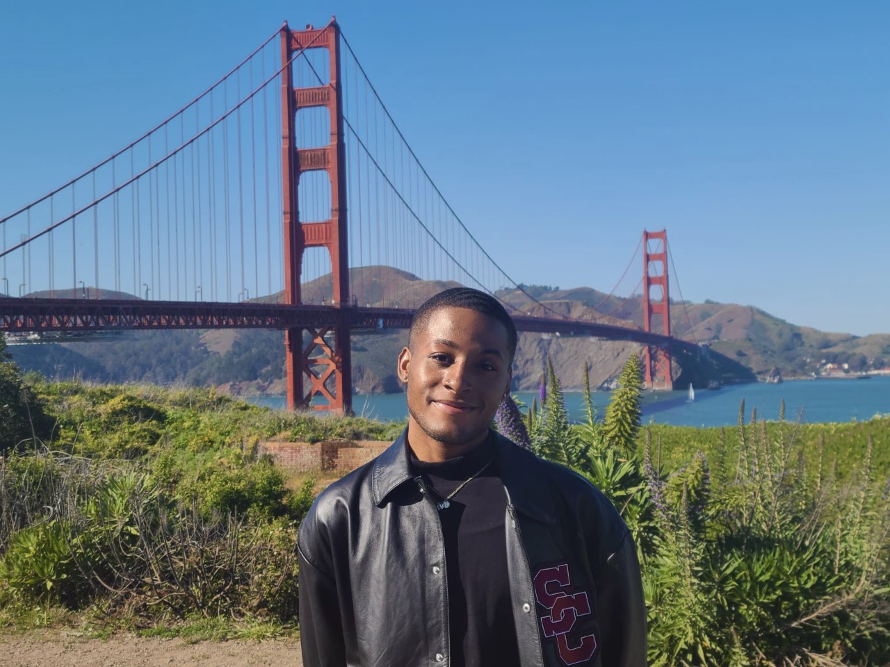 A person with short hair stands in front of the Golden Gate Bridge. They are smiling and wearing a leather jacket.