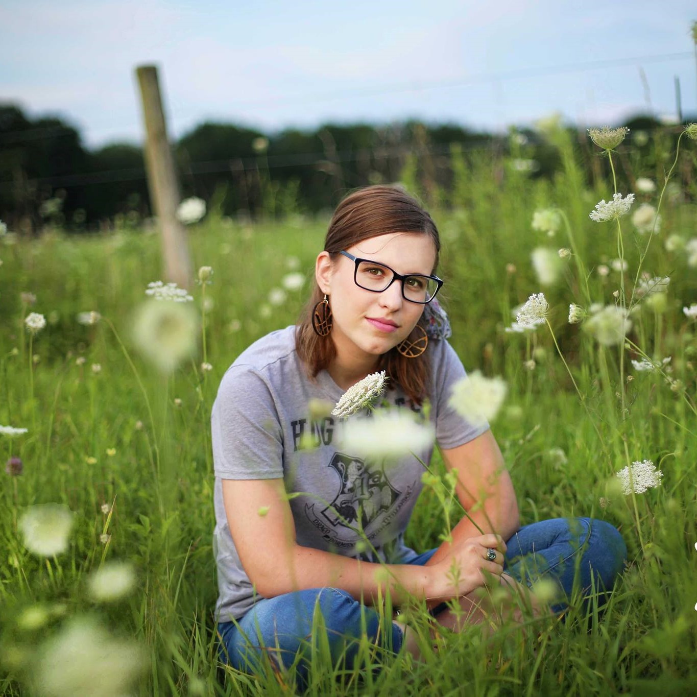 A woman sits in tall grass and flowers. She is wearing glasses and a gray tshirt.