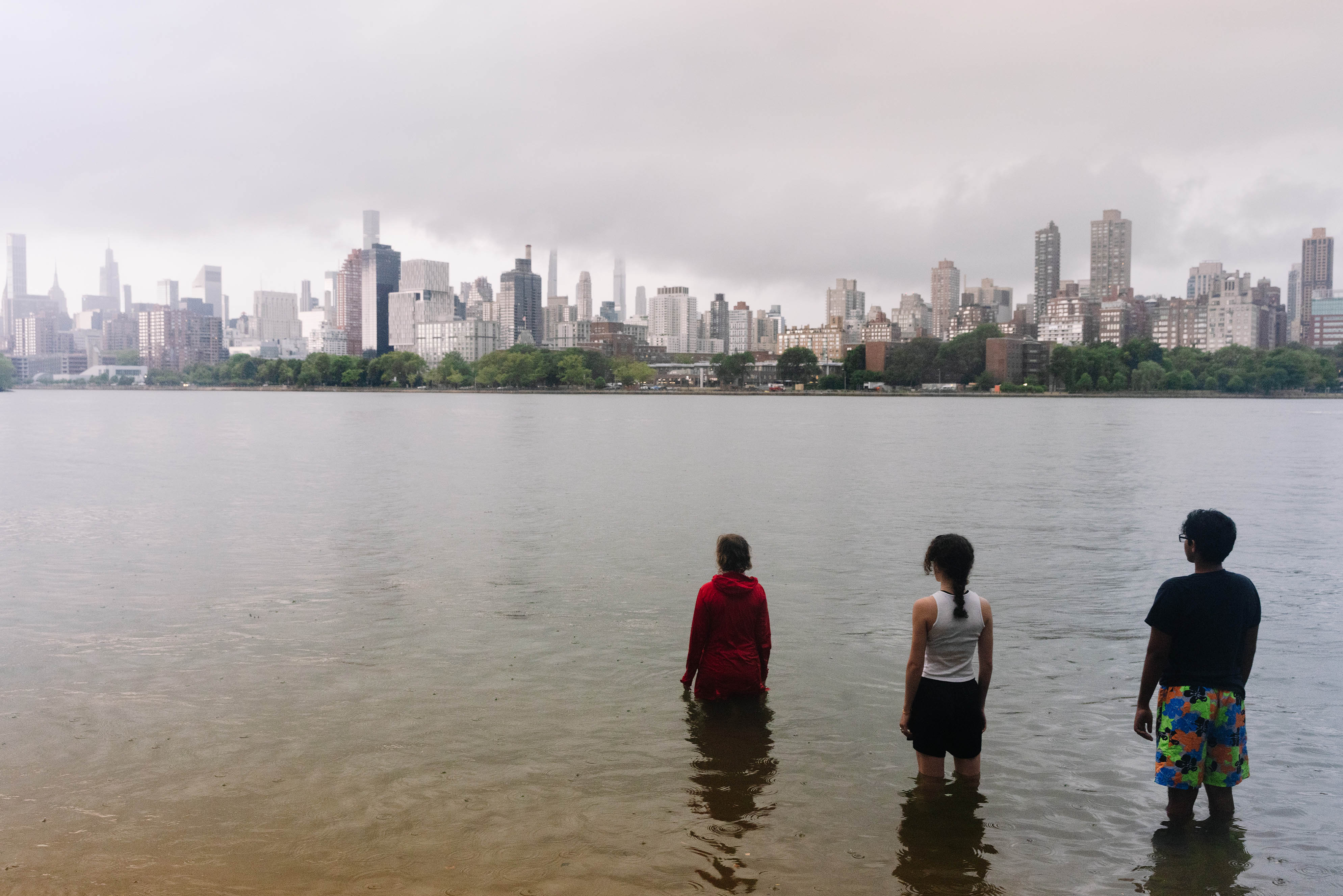 three people stand in the East River, facing the Manhattan skyline, with their backs to the viewer. 