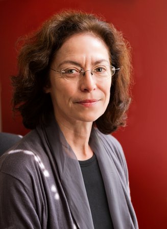 Dr. Di Chiro sits for a portrait wearing wire-rimmed glasses and gray clothing in front of a red backdrop.