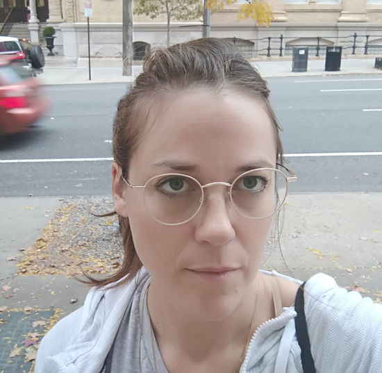 Elisheva is standing on the street, looking slightly up to the camera. She has long hair pulled back and wire rimmed glasses.