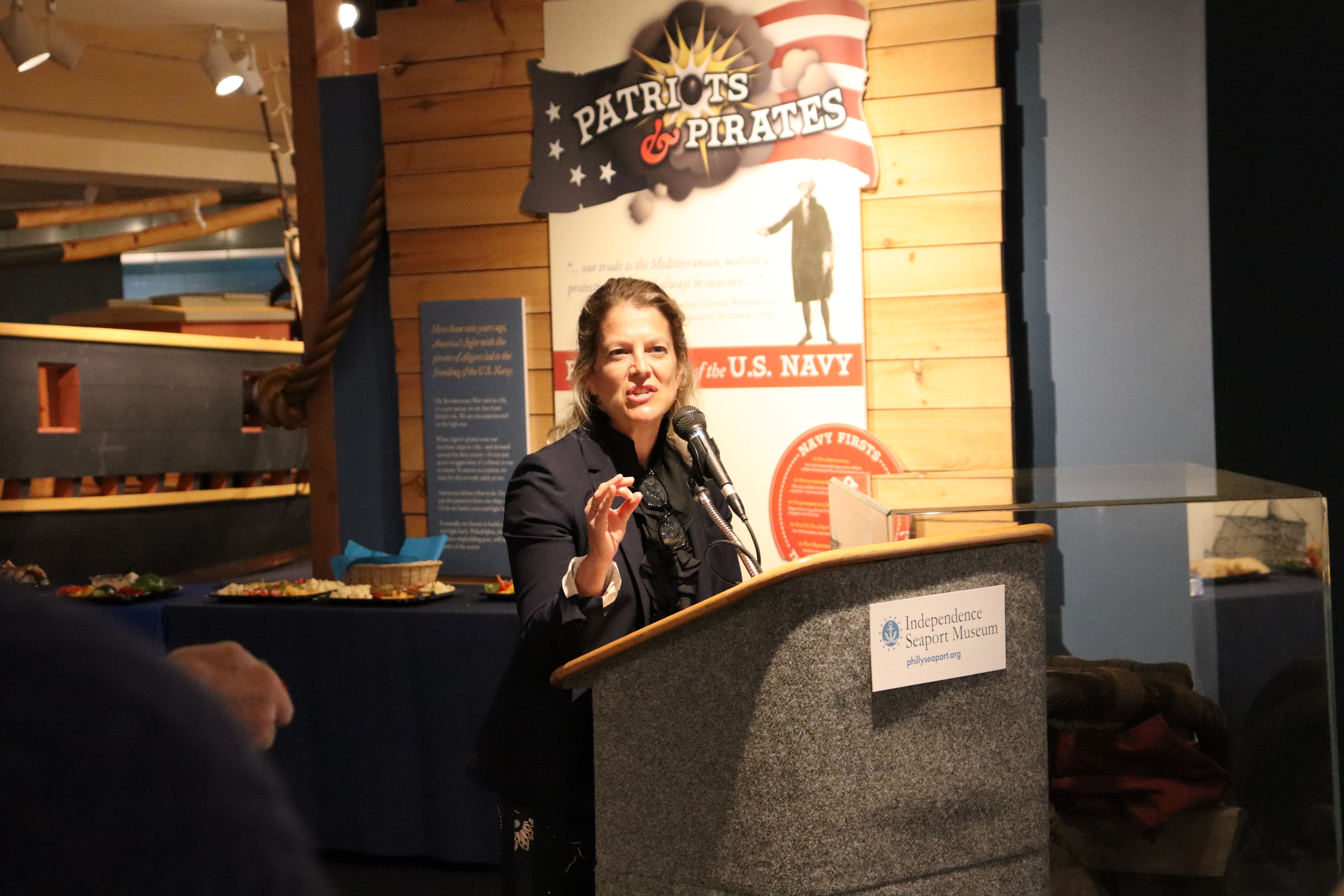 A woman wearing a black blazer with her hair pulled back gives remarks at a podium in a museum gallery.