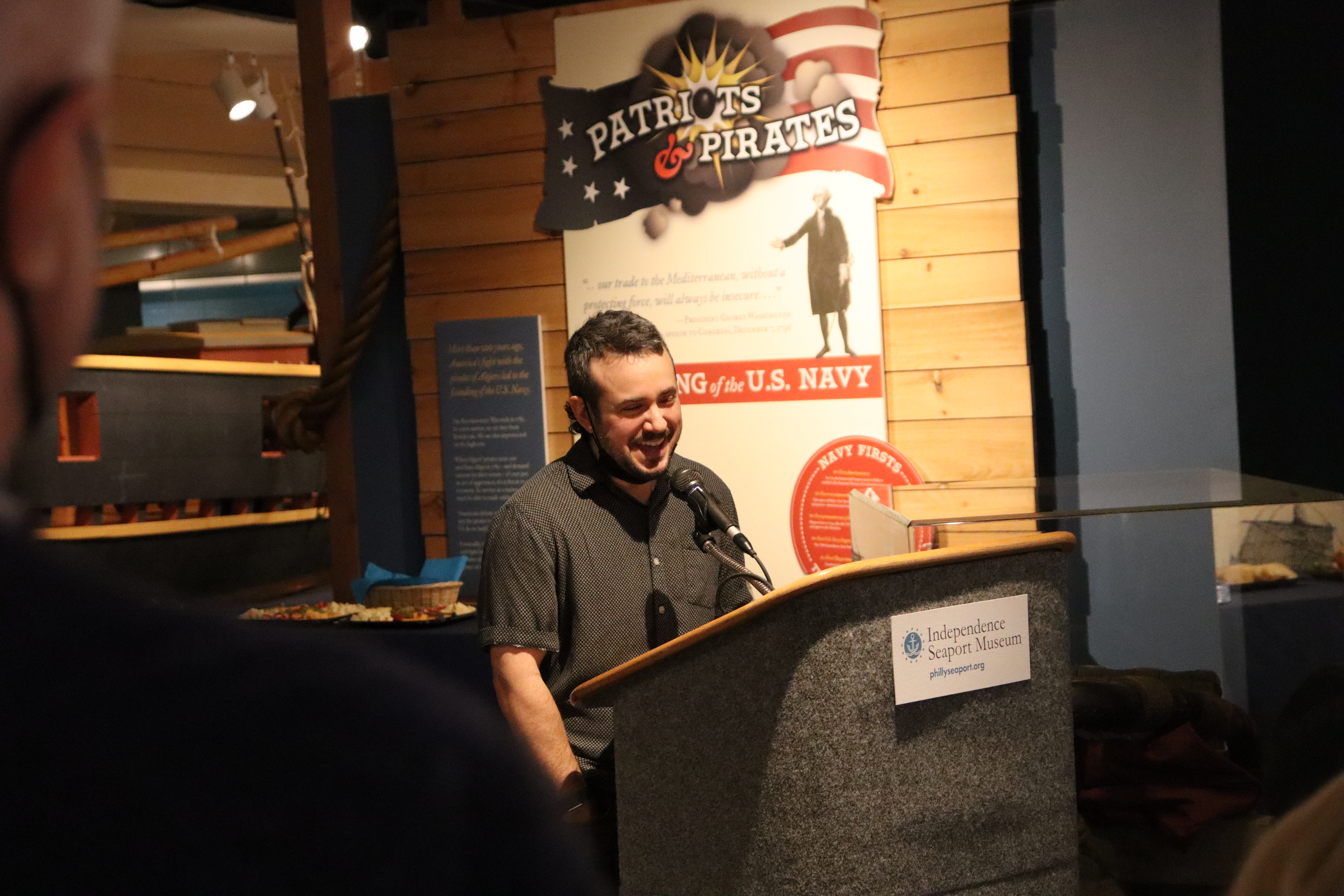 A man in a shirt with small white dots laughs as he gives remarks at a podium in a museum gallery.