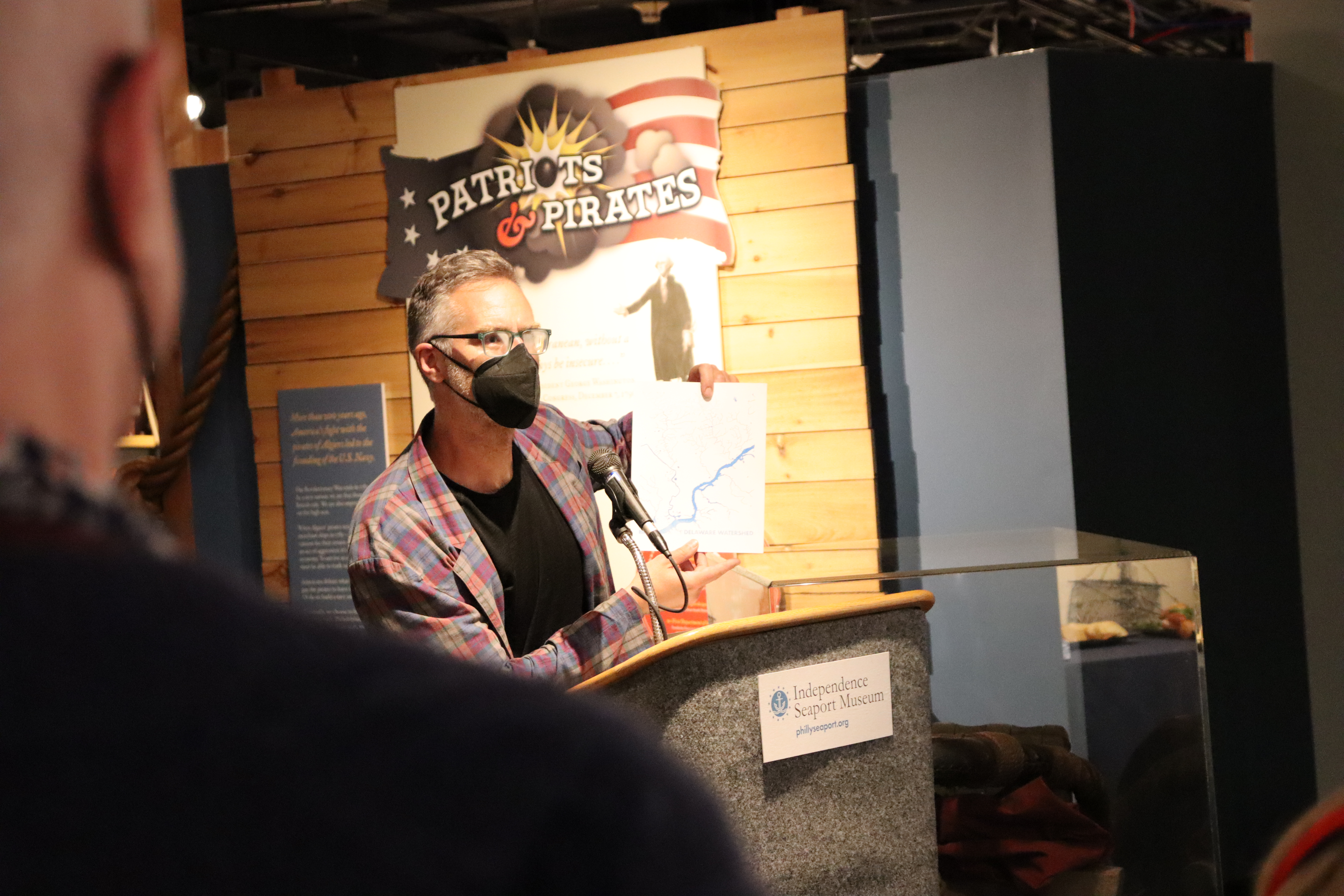 A man in a black face mask holds up a map of a river while giving remarks at podium in a museum gallery.