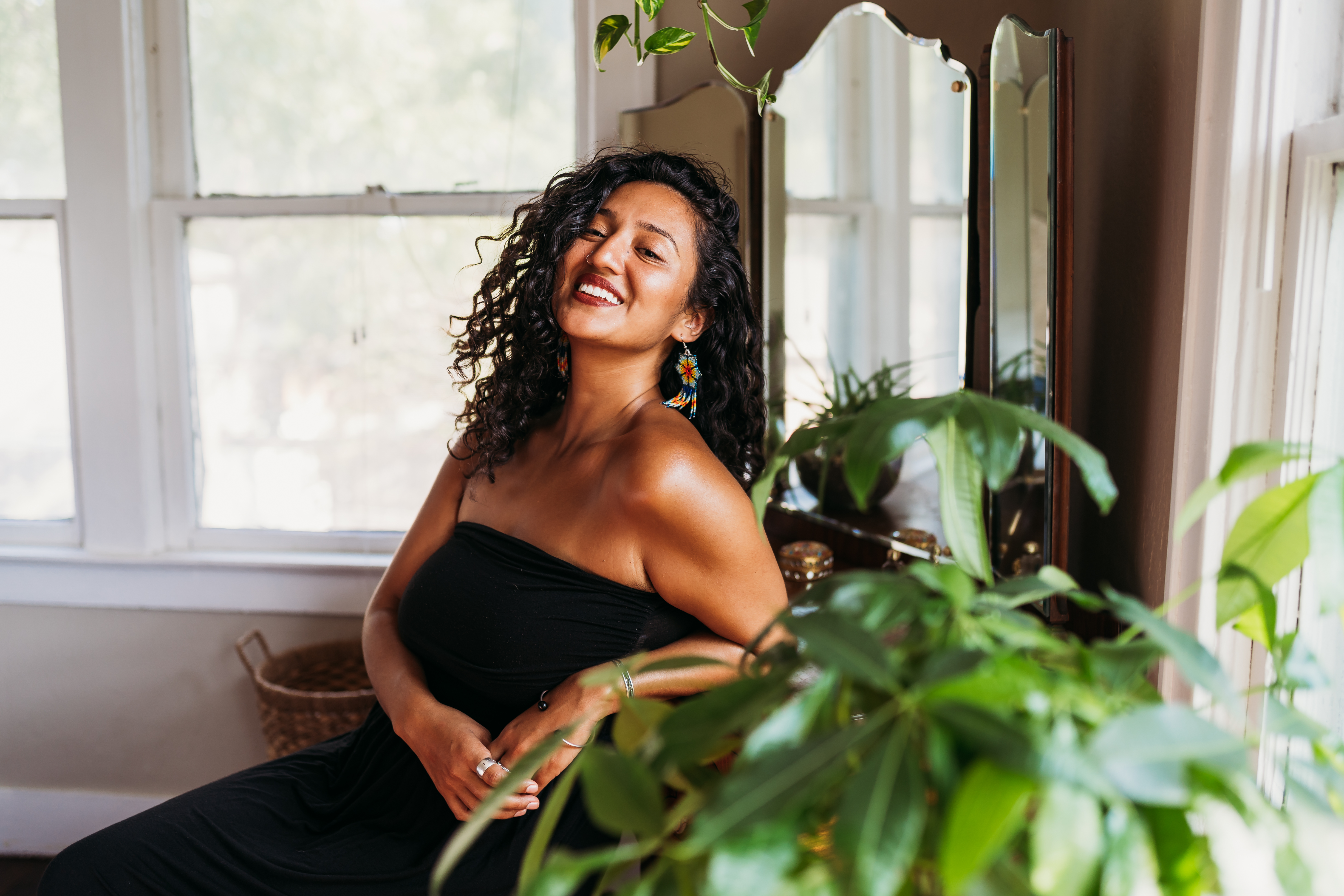 A woman with curly hair in a black strapless dress sits in a chair next to a green plant and smiles at the camera.