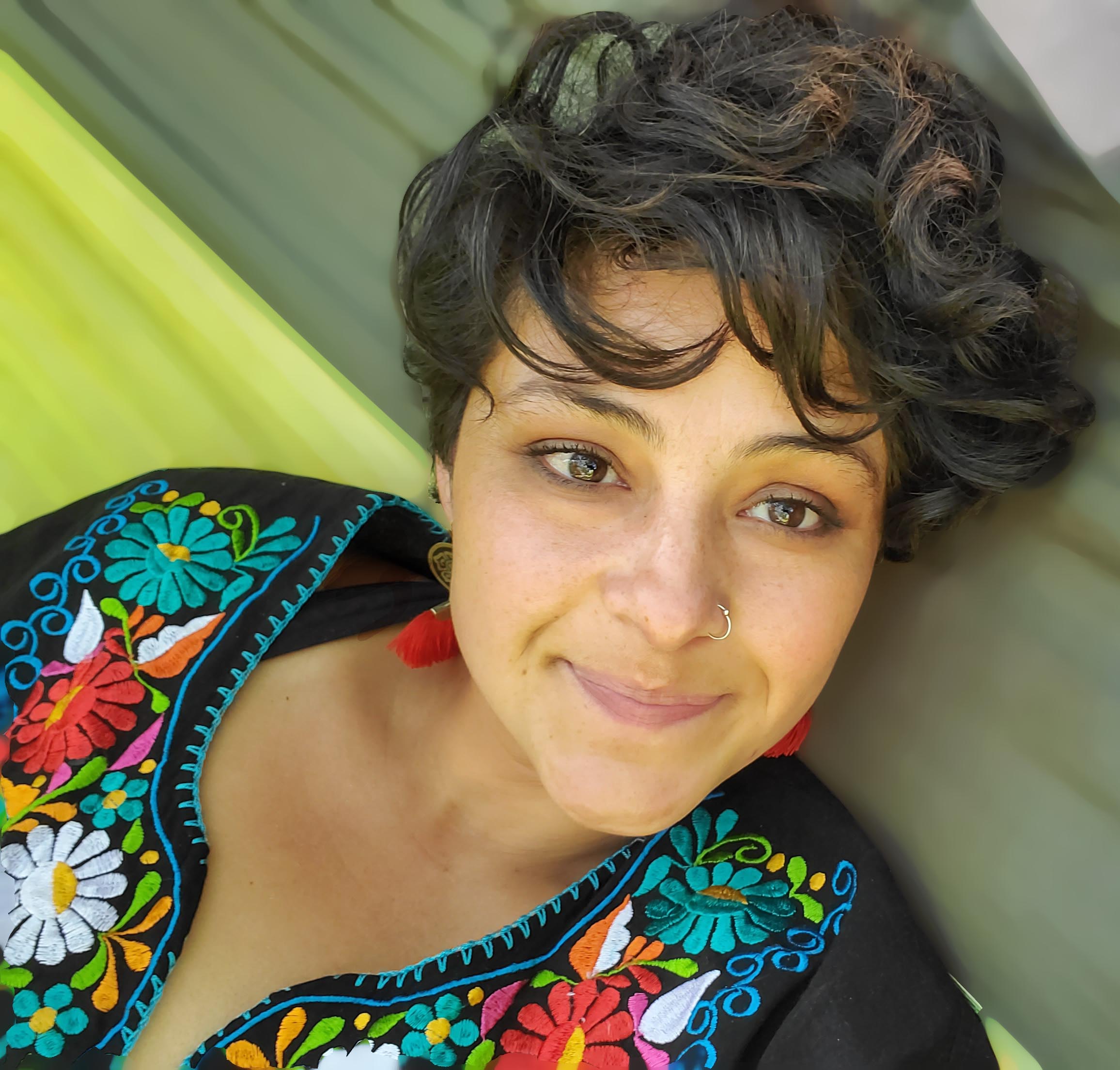 A woman with short curly hair, an embroidered floral dress, lays on a green background and smiles slightly at the camera.