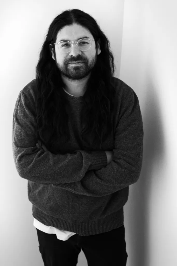 A black and white photo of a man with long hair and a beard, his arms are crossed as he smiles slightly and looks into the camera.