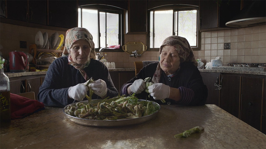 Two old women in headscarves use paring knives to prepare a batch of wild herbs.