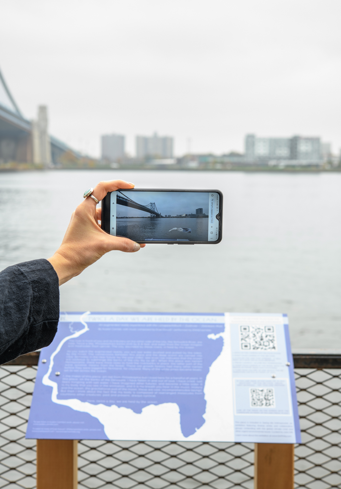 A hand holds a phone up to experience an AR animation of the Delaware River.