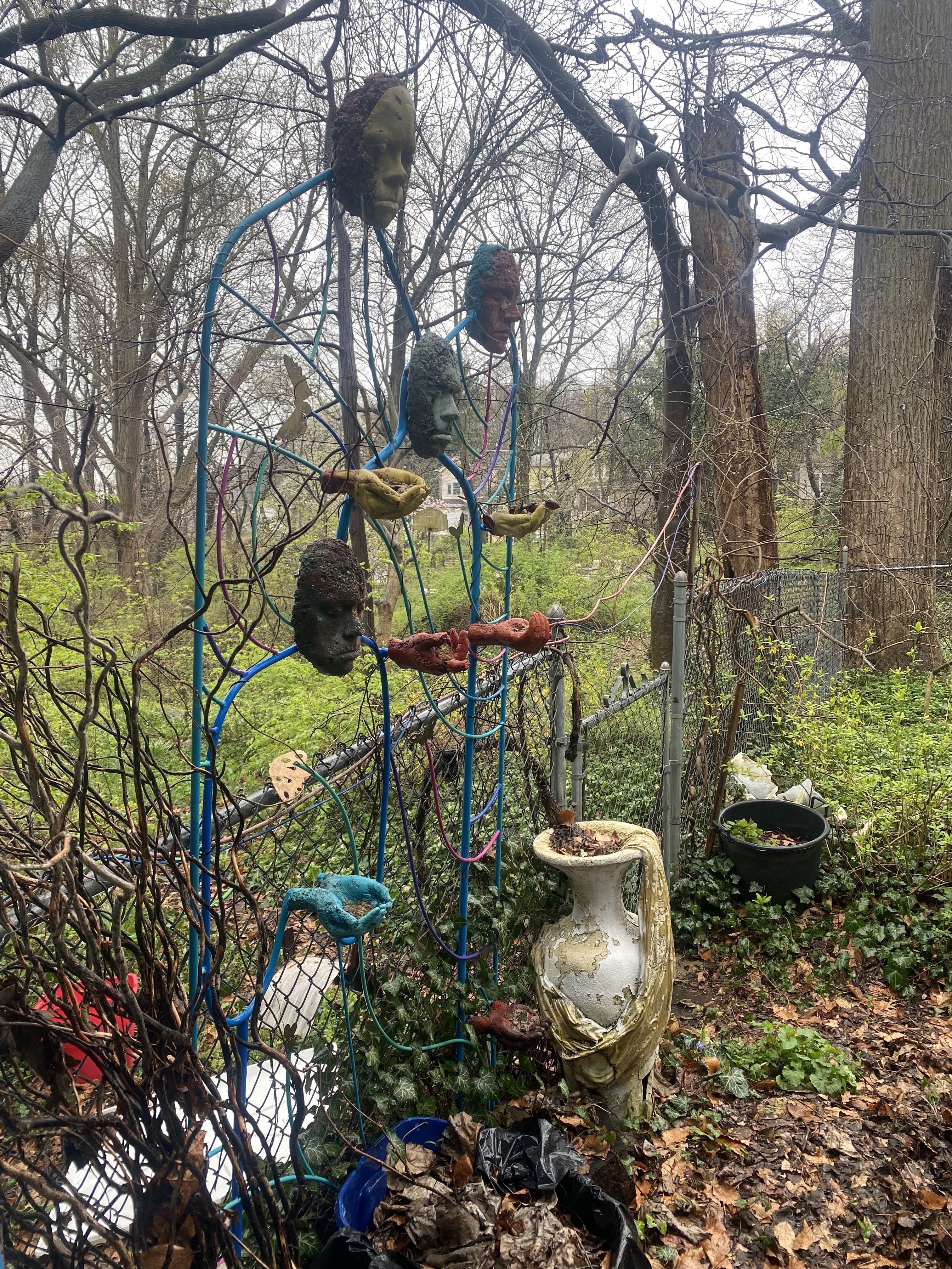 Sculptural human faces and hands are arranged vertically on a trellis in a damp garden.