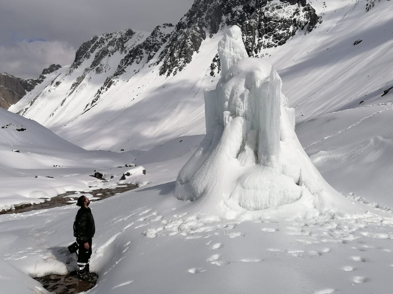 A person in a black winter outfit stands in a snowy mountain scene looking up at a glacial formation
