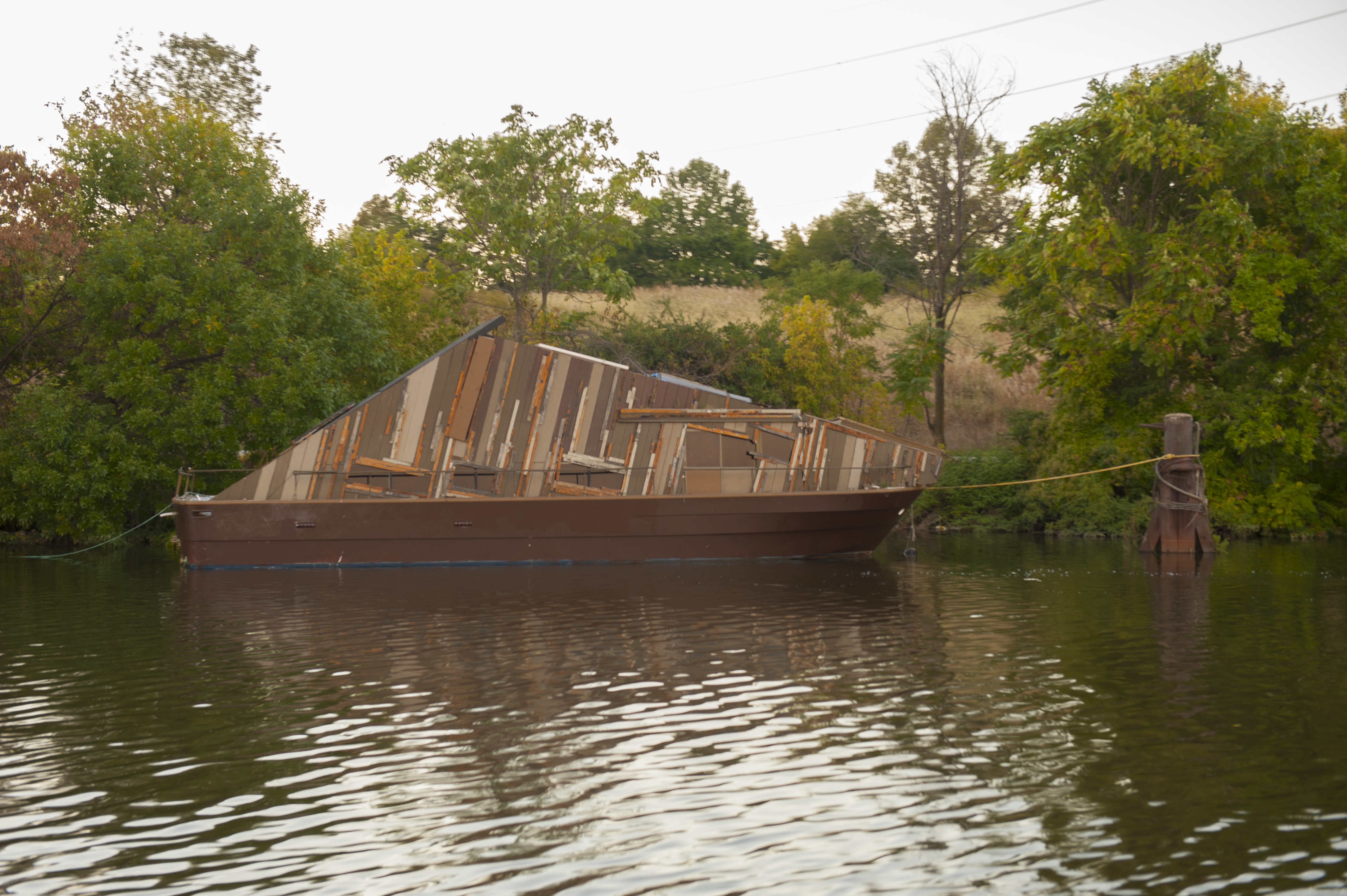 A brown boat floats on a river, tied to a pier. A green riverbank is behind the boat.