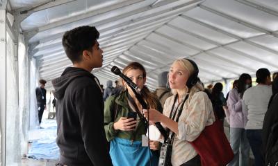 Under a white tent, a student talks into a microphone held by a reporter with headphones 