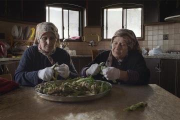 Two old women in headscarves use paring knives to prepare a batch of wild herbs.