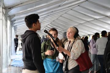 Under a white tent, a student talks into a microphone held by a reporter with headphones 