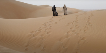Two cloaked figures walk across sand dunes. 